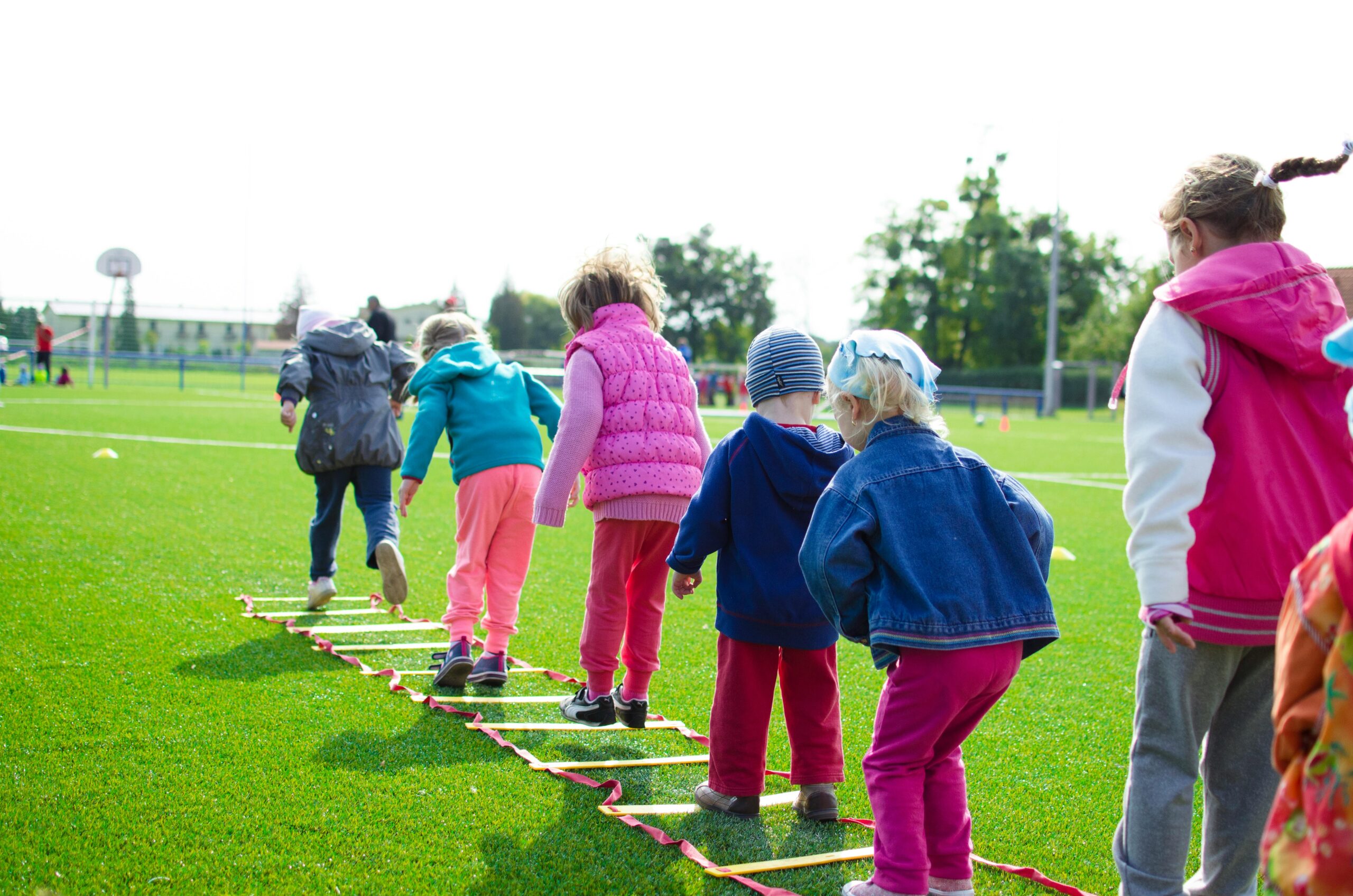 Home Children enjoy an outdoor activity on a grassy field, stepping over a ladder.